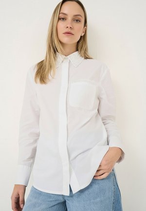 Jeune femme portant une chemise blanche boutonnée avec un col orné de perles et un jean bleu clair, debout avec une main dans la poche.