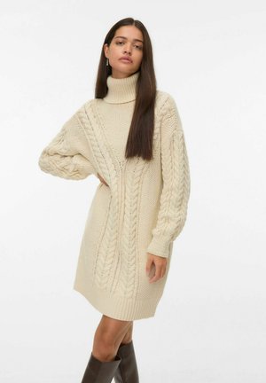Žena v krémových svetrkách s pleteným vzorem a turtleneck šaty, stojící s jednou rukou na boku a tmavými vysokými kozačkami.