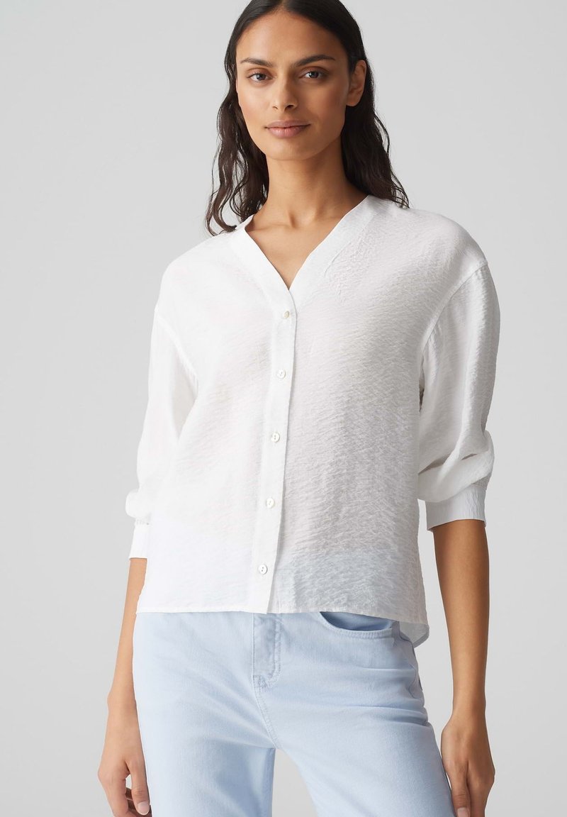 Weißes Hemd mit V-Ausschnitt, langen Ärmeln, strukturiertem Stoff, abgerundeten Säumen und ohne sichtbare Muster, kombiniert mit hellblauen Hosen.