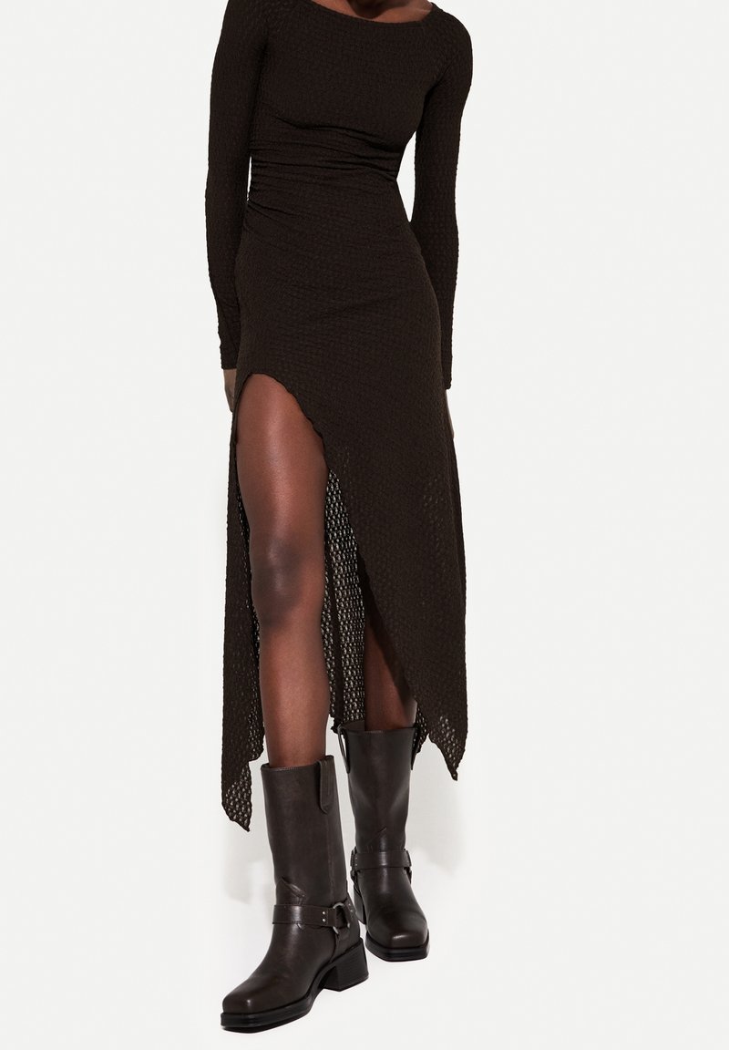 Personne portant une robe noire à manches longues avec une fente haute sur le côté, associée à des bottes en cuir noires mi-mollet avec des boucles.
