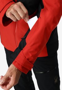 Veste imperméable rouge avec des accents noirs, dotée d'une poche latérale. Une main tire une carte blanche de la poche sur la manche. Tissu texturé.