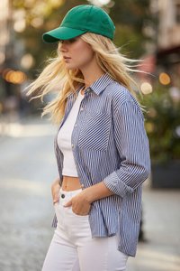 Joven mujer con cabello largo y rubio que lleva una gorra verde, una camisa de rayas azules y blancas sobre un top corto blanco y pantalones blancos en un entorno urbano.
