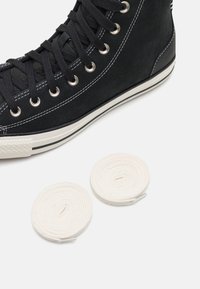Zapatilla alta negra con costuras y suela blanca al lado de dos bobinas de cordones blancos sobre un fondo blanco.