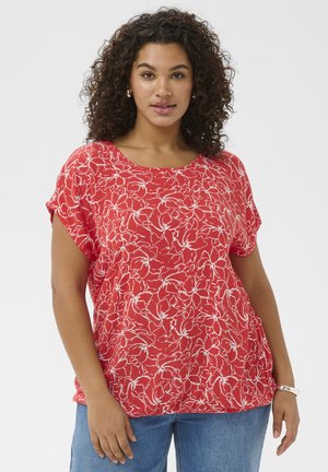 Femeie cu păr creț, purtând o bluză roșie cu flori și mâneci scurte și blugi albaștri, stând cu o mână relaxată și cealaltă ușor ridicată.