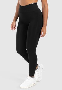 Zwarte legging met een hoge elastische tailleband. Gemaakt van een gladde, rekbare stof. Gecombineerd met witte sportsneakers met een logo.