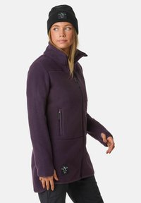 Lila Fleecejacke mit hohem Kragen, Reißverschlusstaschen und einem Logo-Patch. Der Stoff hat eine weiche, strukturierte Oberfläche.