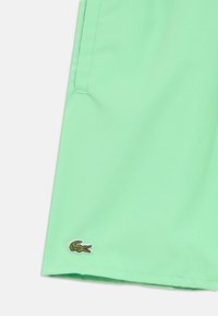 Zöld fürdőnadrág sima textúrával, oldalsó varrásos zsebekkel és a szegélyen egy kis hímzett krokodil logóval.