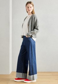 Grauer Strickcardigan, weißes T-Shirt, blaue weit geschnittene Jeans mit rohem Saum und rote Turnschuhe. Naturholzboden und hellgrauer Hintergrund.
