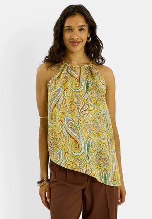 Mujer con top halter amarillo sin mangas con estampado paisley y escote en forma de ojo de cerradura, pantalones marrones, sonriendo, con pulseras y pendientes de aro.