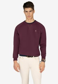 Sudadera burdeos con cuello redondo y puños de canalé, con un pequeño logotipo en el pecho. Combinada con pantalones blancos ajustados.