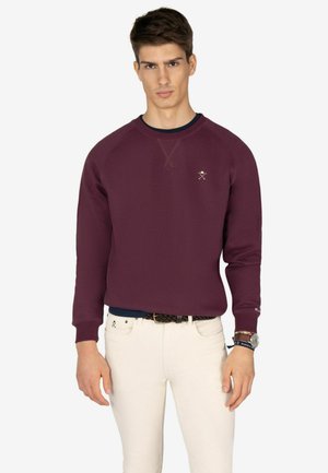 Sudadera burdeos con cuello redondo y puños de canalé, con un pequeño logotipo en el pecho. Combinada con pantalones blancos ajustados.