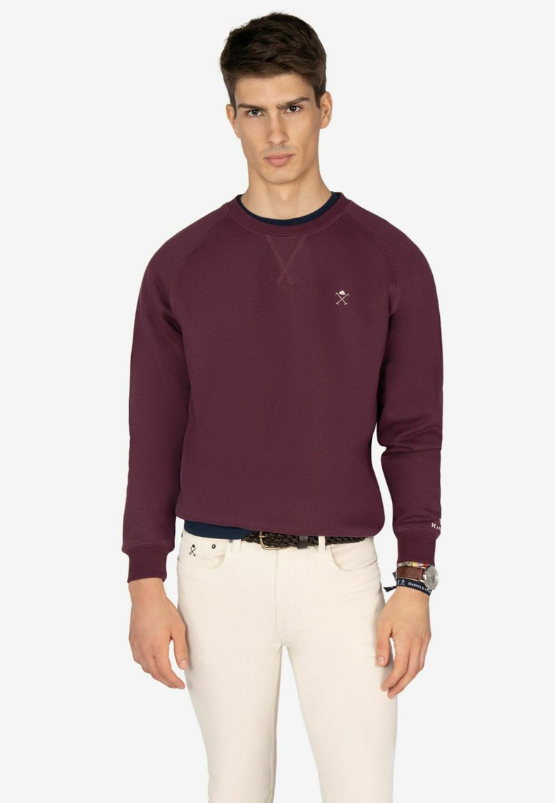 Sudadera burdeos con cuello redondo y puños de canalé, con un pequeño logotipo en el pecho. Combinada con pantalones blancos ajustados.