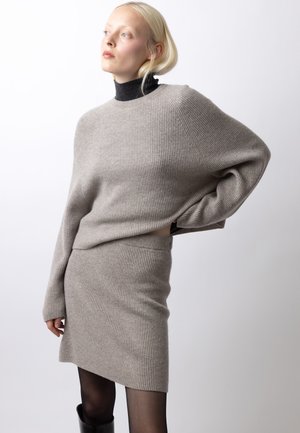Blond kvinde iført lysegrå ribstrikket sweater og matchende nederdel, sort rullekrave under, sorte strømpebukser og sorte støvler, poserer fra siden.
