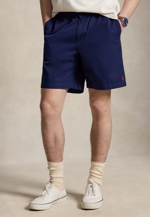 Homme portant un short bleu marine, des chaussettes beige, des baskets blanches et une montre-bracelet, debout les mains dans les poches sur un fond uni.