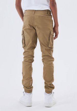 Person iført beige cargobukser med baglommer og elastiske manchetter, kombineret med hvide sneakers og en hvid t-shirt, stående med fronten væk fra kameraet.