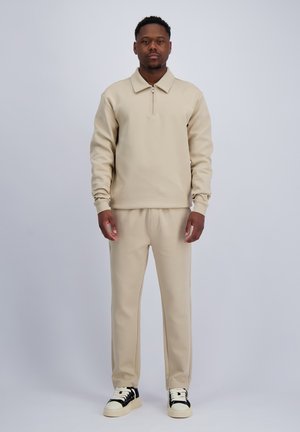 Mann steht und trägt ein beiges Sweatshirt mit Reißverschluss, passende beige Hose und schwarz-weiße Turnschuhe vor hellgrauem Hintergrund.