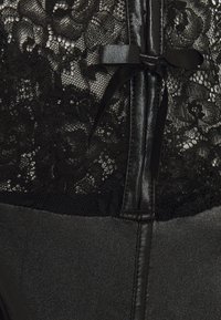 Czarny koronkowy bodysuit z kwiatowym wzorem, z satynową kokardką i nieprzezroczystą dolną częścią. Posiada przezroczystą górę i gładką teksturę całości.