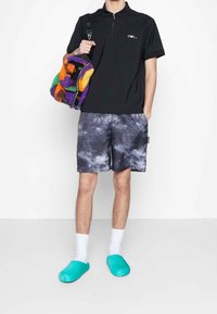 Černé polokošile na zip, šortky s galaktickým vzorem, azurové chlupaté pantofle a barevná texturovaná kabelka přes rameno.