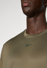 Olive zöld sportpóló sima, matt felülettel, sötétzöld, diszkrét logóval a mellkason, középen elhelyezve, és kerek nyakkivágással.