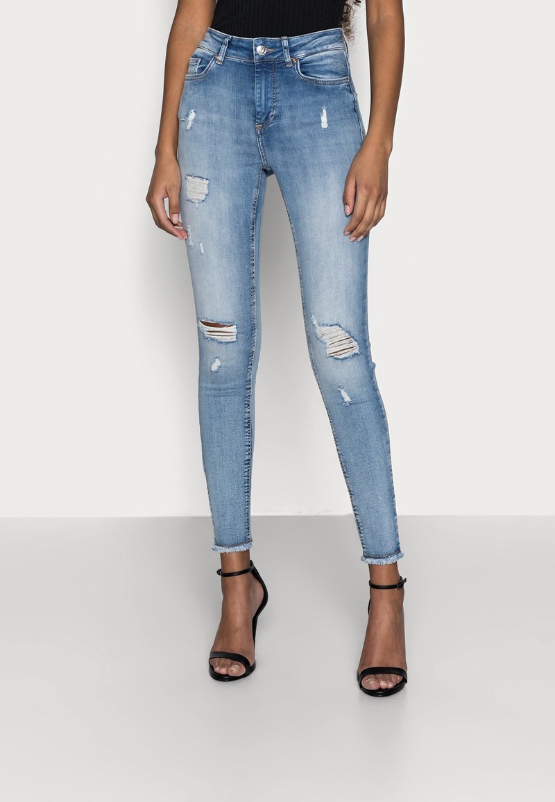Femme portant un jean skinny bleu clair usé et des sandales noires à talons hauts, debout devant un fond blanc uni.
