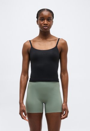 Mladá žena so spletenými vlasmi, ktorá má na sebe čierne tielko a olivovo zelené priliehavé šortky, stojaca na jednoduchom svetlom pozadí.