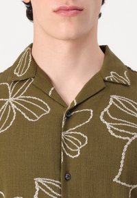 Short-sleeve olive green shirt with white leaf-like embroidery, a notch collar, and black buttons, made from a textured fabric.