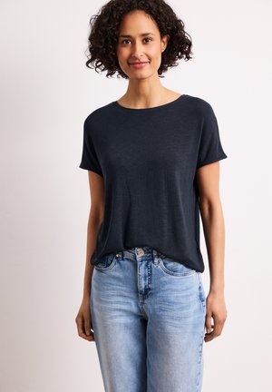 Femme aux cheveux bouclés portant un t-shirt sombre à manches courtes rentré dans un jean bleu clair, debout devant un fond clair uni.