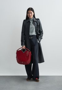Manteau trench en cuir noir, col roulé gris, pantalon bleu marine et sac à main rouge à la forme unique. Chaussures marron avec une semelle texturée.
