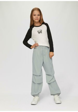 Młoda dziewczyna z długimi włosami stoi z ręką na biodrze, ubrana w białą i czarną koszulę z długim rękawem z motywem motyla, szare spodnie i białe trampki.