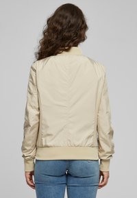 Veste bomber beige à texture lisse, poignets et ourlet côtelés. Dotée d'un col bas et de panneaux latéraux en mesh subtils.
