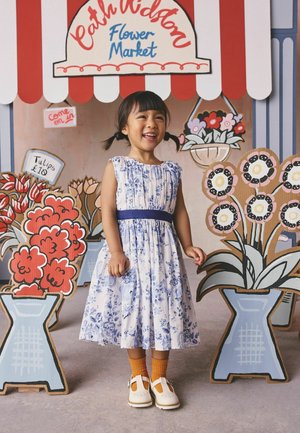 Jauna meitene ar bizītēm, ziedu raksta kleitā un oranžās zeķēs, smaidot stāv starp kartona ziedu izstādēm Cath Kidston ziedu tirgū.