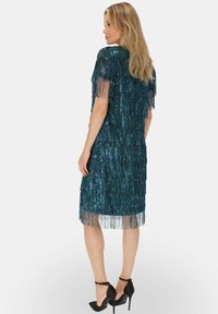 Turkusowa sukienka z cekinami i frędzlami, do kolan, z krótkimi rękawami i prostym krojem. W zestawie z czarnymi szpilkami.