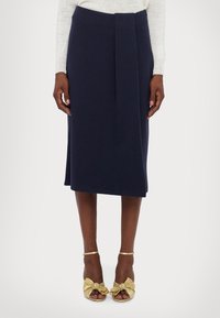 Jupe crayon bleu marine avec un pli devant, confectionnée dans un tissu doux. Associée à des sandales à talons dorées avec des accents de nœud.