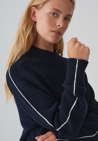 Pull en tricot navy avec un col côtelé haut et des accents rayés blancs le long des manches. Il présente une coupe ample et un tissu texturé.