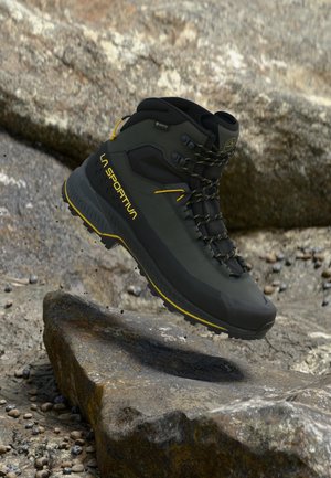 Bocanc de drumeție verde închis și negru cu accente galbene, marca „La Sportiva”, surprins în aer deasupra unui teren stâncos.