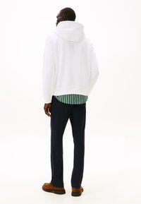 Man facing away wearing a white hoodie, green and white striped shirt, black pants, and brown suede shoes on a white background.