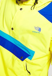 Giacca leggera gialla con accenti blu e turchese. Presenta bottoni a pressione, cappuccio con coulisse e una tasca frontale con patta.