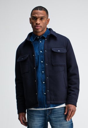 Navy blue wool blend jacket featuring two chest pockets, snap buttons, and a soft texture, layered over a denim shirt. Casual design.
