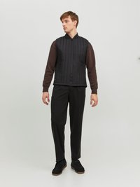 Gilet matelassé noir avec manches côtelées marron, fermeture éclair à l'avant et col montant, associé à un pantalon noir et des chaussures foncées.