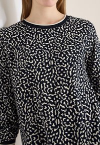 Femme portant un haut à manches longues à motif abstrait noir et blanc avec un col rond, sur un fond uni.