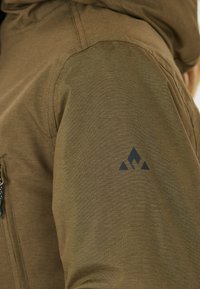 Brun utomhusjacka med texturerad tyg, justerbar huva och en svart logotyp på ärmen. En ficka med dragkedja synlig på sidan.