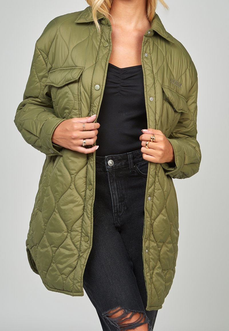 Chaqueta acolchada verde oliva con botones a presión, dos bolsillos frontales y un corte relajado, llevada sobre una blusa negra y unos jeans negros desgastados.