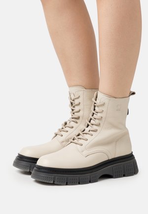 Beige læderstøvler med snøre, tekstureret finish, høj ankeldækning og kraftig sort gummisål med gittermønster.