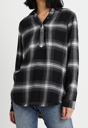 Kobieta ubrana w czarno-szarą koszulę w kratę z długim rękawem, zapinaną na guziki, z postrzępionym dołem, w połączeniu z niebieskimi dżinsami.