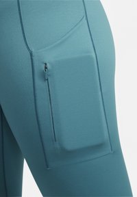 Turkusowe legginsy z elastycznego materiału z kieszonką boczną na zamek, płaskimi szwami i gładką, odprowadzającą wilgoć teksturą tkaniny.