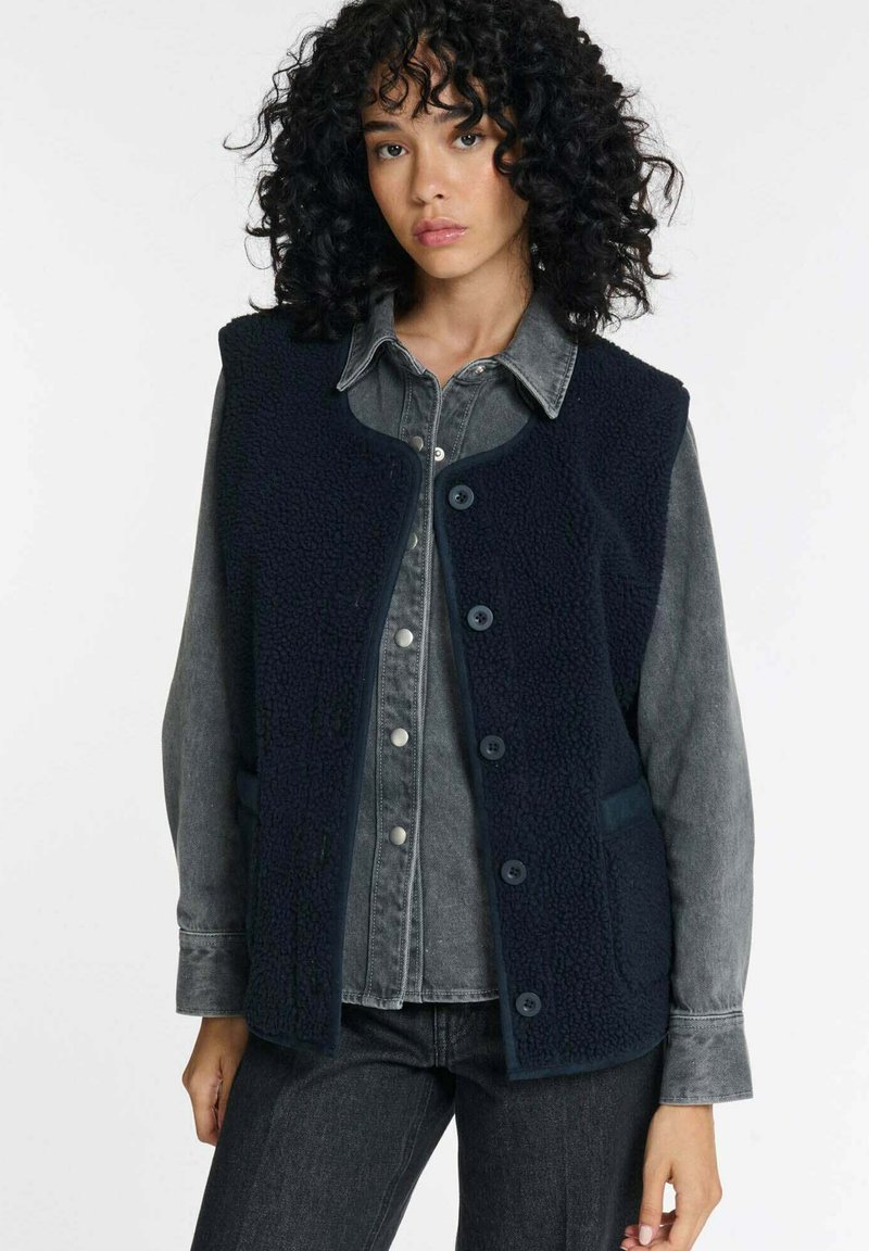 Marineblaue Fleece-Weste mit strukturierter Oberfläche, rundem Ausschnitt, vorderen Knöpfen und Seitentaschen, getragen über einem grauen Jeanshemd.