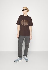 Brun t-shirt med fed beige skrift, grå cargo-bukser med sidelommer og lyseblå sneakers. Enkelt, afslappet fit design.