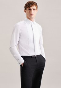 Camicia bianca a maniche lunghe con colletto rigido, caratterizzata da una parte anteriore plissettata e gemelli neri, abbinata a pantaloni neri. Texture del tessuto liscia.