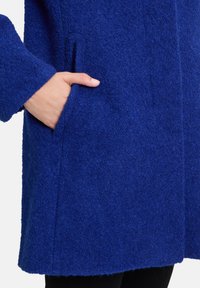 Manteau texturé bleu en matériau doux et pelucheux. Doté d'une poche latérale et d'un design simple. Couture visible sur toute la longueur du manteau.