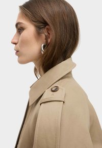 Beige trenchcoat met een gestructureerde kraag, schouderepauletten en een bruine knoopdetail. Glad materiaal met een getailleerde pasvorm.